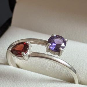 Könnte beinhalten: Ein silberner Bypass-Ring mit zwei verschiedenen, in Krappen gefassten Edelsteinen. Ein Stein ist ein tiefroter, dreieckig geschliffener Granat, der andere ein rund geschliffener, leuchtend violetter Amethyst. Die Ringschiene ist glatt und poliert.