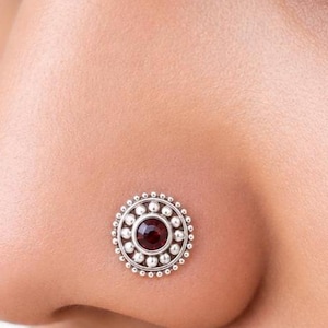 May include: A circular silver nose stud featuring a central dark red gemstone surrounded by a decorative border of small silver beads. The jewelry piece is positioned on the side of a nose, showcasing its intricate metalwork and deep red stone.