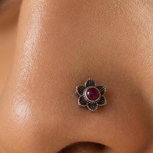 May include: A nose stud featuring a small, oxidized silver flower design with a central, round, deep pink gemstone. The metal petals have a textured, darkened finish that contrasts with the smooth, polished surface of the circular stone.
