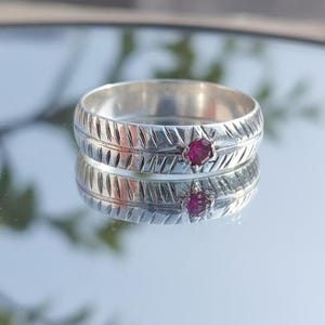 May include: A silver ring with a textured band and a small, round, red gemstone. The ring is displayed on a reflective surface, showcasing its intricate design and the gemstone's vibrant color.