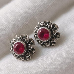 Op de afbeelding: Een paar zilverkleurige oorstekers. Elke oorsteker heeft een centrale, ronde, dieprode edelsteen omgeven door een bloemmotief. De oorbellen zijn geplaatst tegen een witte, getextureerde achtergrond.