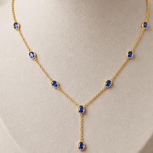 Può includere: Una collana a catena dorata con sette pietre preziose blu dal taglio ovale, distribuite uniformemente e incastonate in castoni dorati. La collana presenta un design a Y con un pendente singolo che pende dal centro.