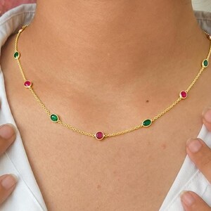 May include: A gold chain necklace featuring evenly spaced, bezel-set round gemstones in alternating red and green colors. The delicate metal chain connects each individual stone setting, creating a simple and symmetrical design around the neck.