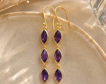 May include: A pair of gold-toned drop earrings featuring three vertically aligned marquise-cut purple gemstones in each earring. The earrings are suspended from gold-toned shepherd hooks and have a slender, elongated design with polished metal settings.