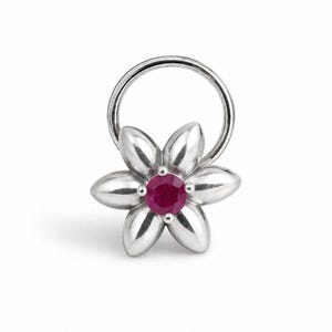 May include: A silver-toned nose ring featuring a six-petaled flower design. The center of the flower is set with a single, round, deep pinkish-red gemstone. The metal petals have a smooth, polished finish and are arranged symmetrically around the stone.