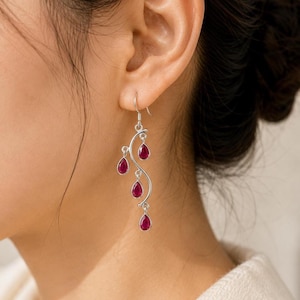 Pode incluir: Um brinco pendente de prata com uma estrutura metálica curva em forma de videira, adornado com quatro pequenas pedras preciosas cor-de-rosa escuro em forma de gota. O brinco está suspenso por um gancho de prata simples.