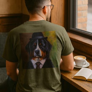 May include: Olive green t-shirt with a portrait of a Bernese Mountain Dog in a suit and hat. The dog's image is on the back of the shirt. A person wearing glasses is sitting at a table with a coffee cup and an open book.