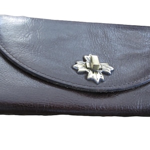 Op de afbeelding: Een donkerbruine leren clutch met een gebogen flap en een zilverkleurige blad-vormige sluiting. De clutch is rechthoekig en lijkt gemaakt van getextureerd leer. De sluiting is gecentreerd op de flap.