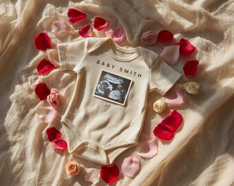 May include: Cream-colored baby onesie with the text "BABY SMITH" and a sonogram image. The onesie is surrounded by red, pink, and cream-colored rose petals and small roses, arranged on a cream-colored fabric background.