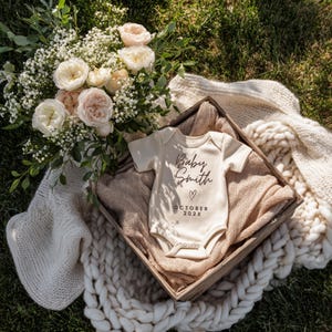 May include: A cream baby onesie with the text "Baby Smith" and "October 202X" in a wooden box. The box is surrounded by a bouquet of white and peach roses, a beige blanket, and a chunky knit blanket.