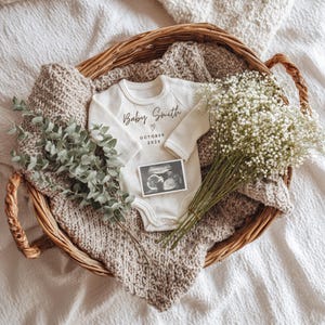 May include: A wicker basket contains a cream baby onesie with "Baby Smith" printed on it, an ultrasound scan, a knitted blanket, eucalyptus, and a bouquet of white flowers. A charming announcement.