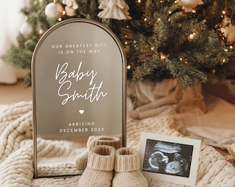 Puede incluir: Un letrero espejado con el texto "OUR GREATEST GIFT IS ON THE WAY" y "Baby Smith" con un símbolo de corazón, y "ARRIVING DECEMBER 202X". Un par de botines de bebé de punto beige y una imagen de ultrasonido frente a un árbol de Navidad.