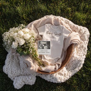 May include: A cream babygrow with the words "Baby Smith" and a heart, in a wicker basket with a blanket and white roses. An ultrasound image reads "Arriving October 202X". The scene is set on green grass.