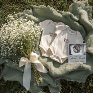Puede incluir: Dos bodies de bebé color crema con las palabras "baby one" y "baby two" en escritura marrón. Una imagen de ultrasonido con el texto "Smith Twins OCTOBER 202X" está al lado de los bodies. Un ramo de flores blancas con una cinta crema está sobre una manta verde.