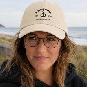 Puede incluir: Una gorra de béisbol de color beige con un gráfico negro de un ancla y el texto WIDOW'S BAE EST. 1681 DEFINITELY NOT CURSED! impreso en la parte delantera. La gorra tiene una visera curva y una corona estructurada con un botón en el centro.