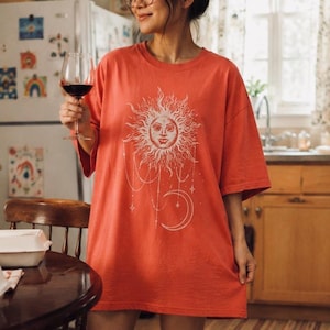 Könnte beinhalten: Ein übergroßes korallenfarbenes T-Shirt mit einem großen weißen Grafikdruck einer Sonne mit menschlichem Gesicht, umgeben von hängenden Sternornamenten und einer Mondsichel. Das T-Shirt hat kurze Ärmel und einen Rundhalsausschnitt.