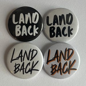May include: Four circular badges with the words "LAND BACK" in a handwritten font. The badges are in black, white, and a combination of white and orange. The badges are on a white surface.