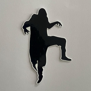 May include: Black silhouette sticker of a person in a dynamic pose, with one leg raised and arms outstretched. The sticker has a white border, creating a contrast against the black figure. The sticker is on a light grey background.