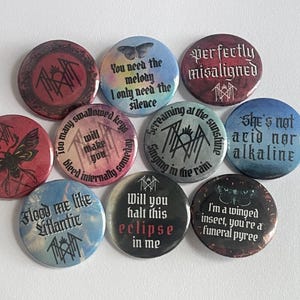 May include: A set of ten circular badges with dark, gothic-inspired designs. The badges feature phrases and illustrations, including a butterfly and insect. The colour palette is predominantly dark, with text in a stylized font.