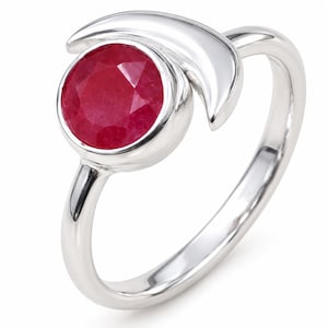 Op de afbeelding: Een ring van gepolijst zilver met een ronde, gefacetteerde, dieprode edelsteen in een zetting. De band bevat een gladde, gebogen halvemaanvorm die gedeeltelijk om de ronde zetting heen loopt, wat zorgt voor een strakke en reflecterende metalen afwerking.
