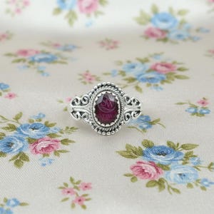 May include: A silver ring with an oval, deep red gemstone. The ring has an ornate, filigree design around the stone and on the band. The gemstone is set in a beaded bezel. The ring is displayed on a floral patterned fabric.