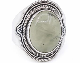 May include: A silver ring featuring a large, oval-shaped, pale green cabochon stone with dark inclusions. The setting is decorated with a twisted rope border and small beaded accents, creating a textured frame around the central gemstone.