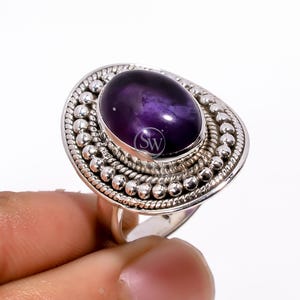 May include: A silver ring with a large, oval, deep purple gemstone. The gemstone is set in a decorative bezel with a beaded border. The ring's band is simple, and the design is elegant. The ring is a statement piece.