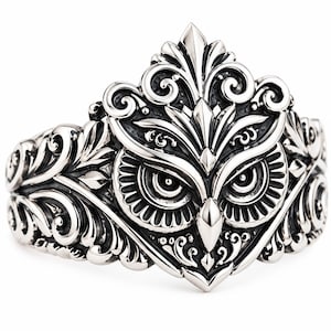 May include: A silver-toned metal ring featuring a detailed owl face at the centre, surrounded by intricate, raised scrollwork and leaf-like patterns that extend across the band. The design has a polished finish with darkened recessed areas.
