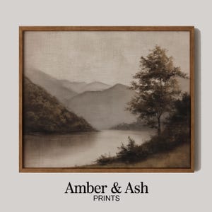 May include: A framed landscape print featuring a serene lake scene with mountains in the background. The artwork is rendered in muted tones of brown and gray, with a tree on the right side. The text "Amber & Ash PRINTS" is at the bottom.