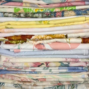 May include: A stack of folded vintage bed sheets. The sheets display a variety of floral patterns in pastel colors, including pink, yellow, blue, and green. The sheets are neatly stacked, showcasing the different designs and textures.