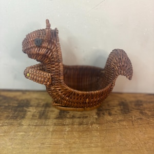 May include: A woven brown squirrel-shaped basket. The basket features a detailed design with a tail, ears, and a small eye. The basket is made of woven material and is designed to hold small items. The squirrel basket is a decorative item.