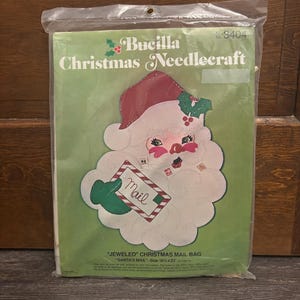May include: A Bucilla Christmas Needlecraft kit in a plastic package. The kit features a Santa Claus design holding a mail bag that reads "Mail". The bag measures 18.5 x 23 inches.