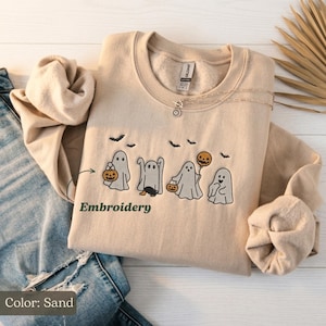 Peut inclure: Un sweat-shirt à col rond couleur sable avec une broderie représentant quatre fantômes. Les fantômes sont illustrés dans diverses poses, certains tenant des citrouilles ou des ballons, avec de petites chauves-souris noires brodées au-dessus.
