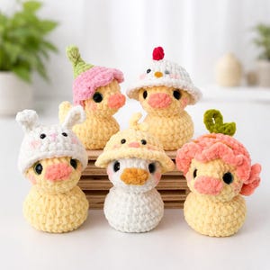 May include: A collection of five crocheted duck figurines, each wearing a different hat. The ducks are primarily yellow, with pink, white, and green accents. The hats include a bunny, a flower, and a chicken.