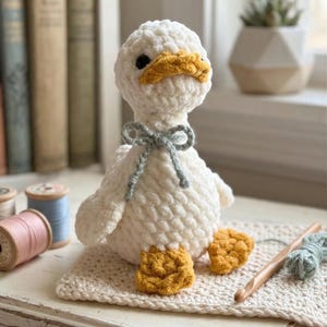 Puede incluir: Un pato blanco hecho a mano con ganchillo, con pico y patas amarillas, y una pajarita gris. El pato está sentado sobre un cuadrado de ganchillo color crema. También se ven un ganchillo de madera y carretes de hilo.