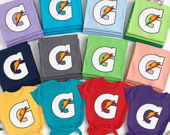 May include: A collection of twelve folded and unfolded t-shirts in various colors including purple, blue, light blue, green, dark gray, yellow, red, and peach. Each shirt features a large white letter G with an orange lightning bolt graphic.