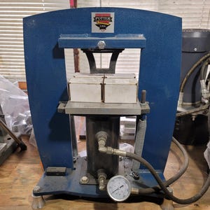 May include: A blue Potter USA hydraulic press featuring a central cylinder, a pressure gauge, and a metal plate supporting four small white rectangular boxes. The machine has a sturdy frame, vertical springs, and visible hydraulic hoses.