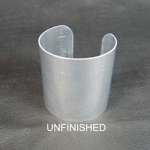 May include: A silver-colored cuff bracelet with a curved design. The open-ended bracelet is cylindrical and has the word "UNFINISHED" printed at the bottom. The cuff is made of a shiny metal.