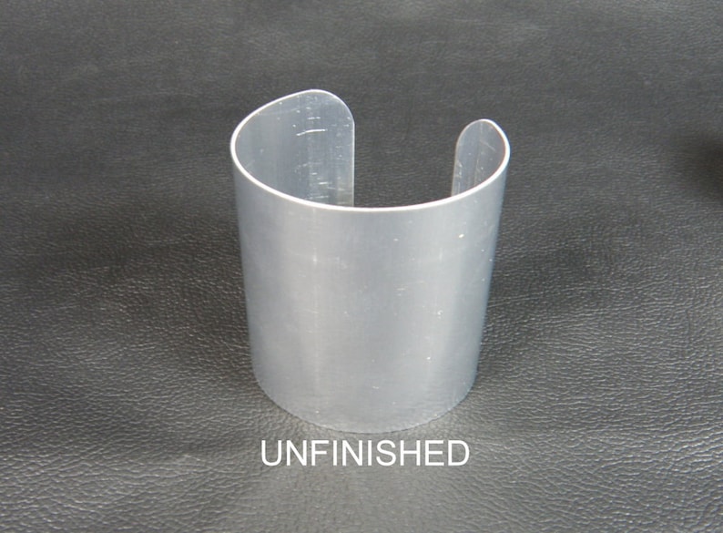 May include: A silver-colored cuff bracelet with a curved design. The open-ended bracelet is cylindrical and has the word "UNFINISHED" printed at the bottom. The cuff is made of a shiny metal.