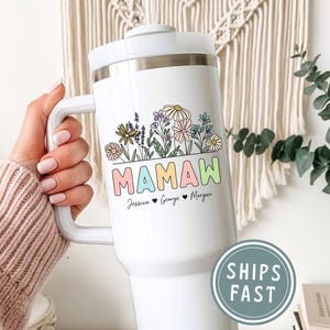 May include: White travel mug with a handle, featuring a floral design above the word "MAMAW" in pastel colors. Below the word are the names Jessica, George, and Morgan. The mug has a white lid and handle.