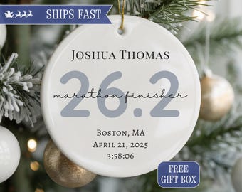 May include: White ceramic Christmas ornament with the text "JOSHUA THOMAS 26.2 marathon finisher" and event details. The ornament includes the date April 21, 2025, and time 3:58:06. The ornament is hanging on a Christmas tree.