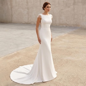 Op de afbeelding: Een vloerlange witte trouwjurk met een boothals, korte mouwen en een aansluitend lijfje. De rug en schouders zijn versierd met kleine witte parels. De rok loopt uit in een lange sleep, wat zorgt voor een strak en elegant silhouet.