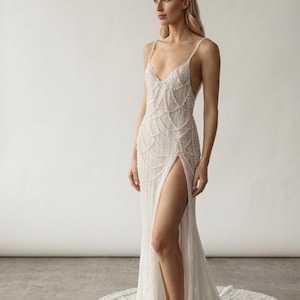 Pode incluir: Um vestido branco longo até ao chão com decote em V profundo, alças finas e um padrão intrincado de missangas verticais e drapeadas. O vestido inclui uma fenda lateral alta que revela a perna e uma cauda longa no chão.