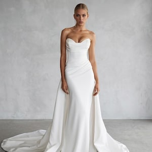 Può includere: Un abito lungo bianco senza spalline caratterizzato da un corpetto strutturato a corsetto con scollo drappeggiato e gonna aderente. L'abito include una sopragonna staccabile che crea un lungo strascico, realizzato in tessuto bianco liscio.
