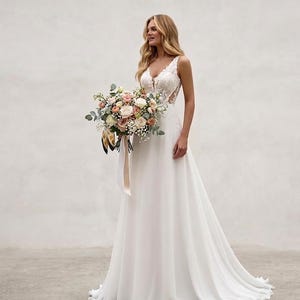 May include: A floor-length white wedding dress featuring a sleeveless bodice with intricate lace detailing, a deep V-neckline, and a flowing skirt. The dress is paired with a large bouquet of mixed pastel flowers tied with a long cream ribbon.