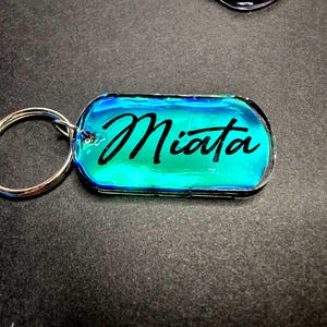 May include: A rectangular keychain with a silver key ring. The keychain has a blue and green iridescent background with the word "Miata" in black script. The keychain is a car accessory.
