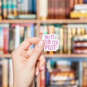May include: A white sticker with the text "DO IT FOR THE PLOT" in pink, held in front of a bookshelf. The sticker has a pink star design. The background is a blurred bookshelf filled with books.