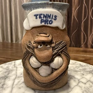 Puede incluir: Taza de cerámica caprichosa con una cara de dibujos animados, sosteniendo tres pelotas de tenis. La taza tiene un borde azul y una pancarta que dice "TENNIS PRO" en letras azules. La taza es principalmente marrón con detalles beige y blancos.