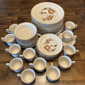 Puede incluir: Un gran juego de vajilla de gres con acabado beige moteado y bordes marrones. La colección incluye pilas de platos llanos y platos de ensalada decorados con motivos florales marrones, junto con numerosas tazas y cuencos a juego.