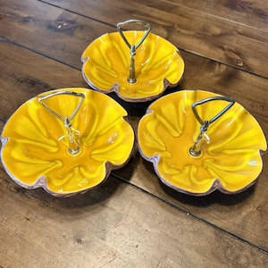 May include: Three yellow ceramic serving dishes with gold-colored handles. The dishes have a ruffled edge and a glossy finish. The dishes are arranged on a wooden surface.